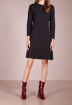 Vrouw in een zwarte jurk tot op de knie met lange mouwen, gecombineerd met glanzende donkerrode enkellaarzen, staande op een gladde vloer.