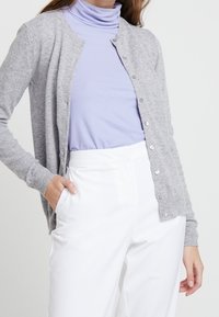 Ljusgrå cardigan med rund halsringning, knappstängning och långa ärmar, lagd över en ljuslila polo, kombinerad med vita byxor.