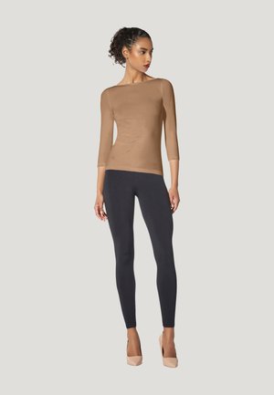 Donna con capelli ricci con una maglia aderente a maniche lunghe color cammello, leggings neri e tacchi nude, in piedi davanti a uno sfondo chiaro e uniforme.