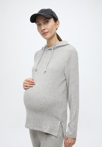 Pelēka grūtnieču kapuce ar aukliņu kapuci, sānu šķēlumiem un mīkstu tekstūru. Valkāta ar melnu cepuri, izceļot apaļo vēderu.