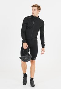 Veste de cyclisme noire avec fermeture éclair, portée sur un short noir. Le mannequin tient un casque noir, présentant des accents réfléchissants et un design ajusté.