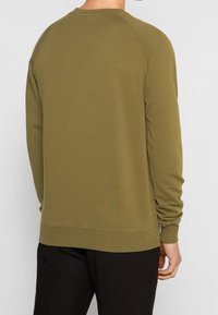 Muž v olivově zelené mikině s dlouhým rukávem a černých kalhotách, zobrazen zezadu na jednobarevném pozadí.