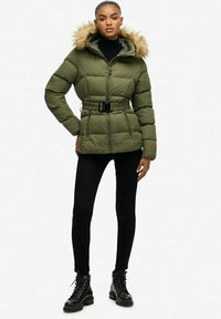 Chaqueta acolchada de color verde oliva con capucha de piel sintética, cinturilla ajustada, cremallera frontal y diseño acolchado. Combinada con leggings negros y botas con cordones.