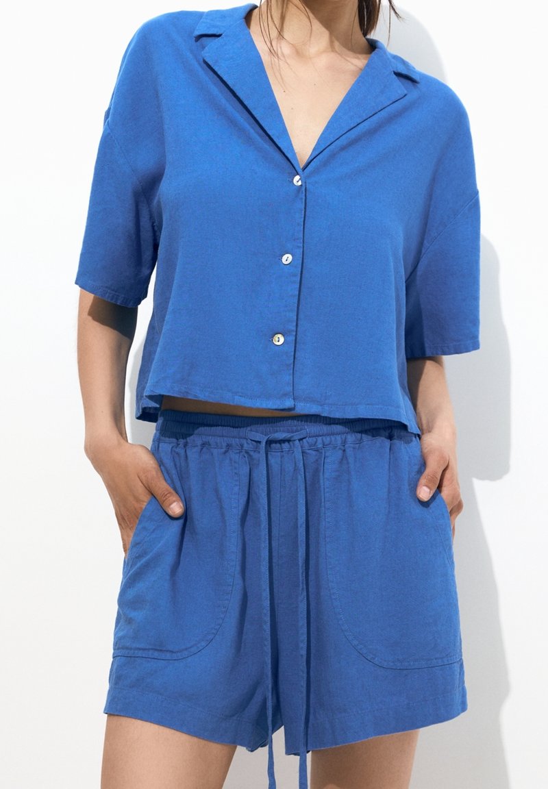 Femme portant une chemise bleue à manches courtes boutonnée et un short assorti à cordon, mains dans les poches, sur fond blanc.