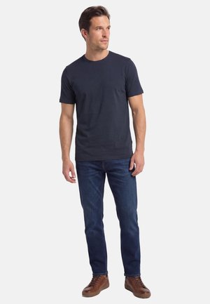 Mand stående iført en mørkeblå t-shirt, blå jeans og brune afslappede sko, ser let mod højre.