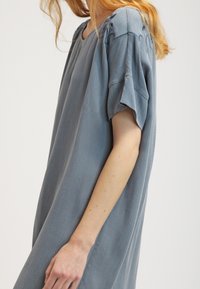 Modré oversized šaty s texturovanou látkou; mají kulatý výstřih a krátké, volné rukávy se dekoračním šitím na ramenou.
