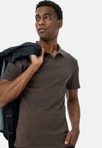 Brun ribbet polo t-shirt med krave, tekstureret stof og korte ærmer, båret med en mørk denimjakke over skulderen.