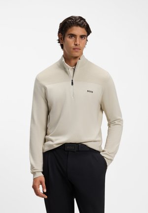 Homme portant un pull beige à zip quart avec le logo "BOSS" et un pantalon noir avec une ceinture assortie, debout devant un fond blanc.