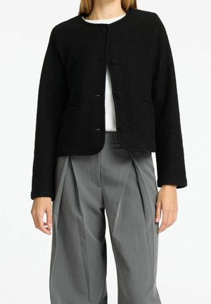 Nainen pukeutunut mustaan villatakkkiin, joka on napitettu valkoisen paidan päällä, ja hänellä on yllään korkeavyötäröiset harmaat laskostetut housut, seisoo valkoista taustaa vasten.