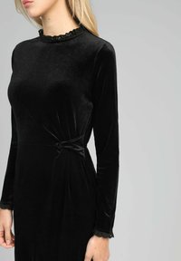 Femme portant une robe noire en velours ajustée avec des manches longues, bordée de dentelle au col et aux poignets, et un détail froncé à la taille.