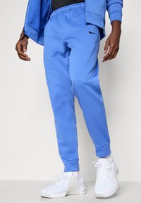 Blue cotton blend pants with an elastic waistband and cuffs, featuring a black Nike logo on the thigh, paired with white athletic shoes.