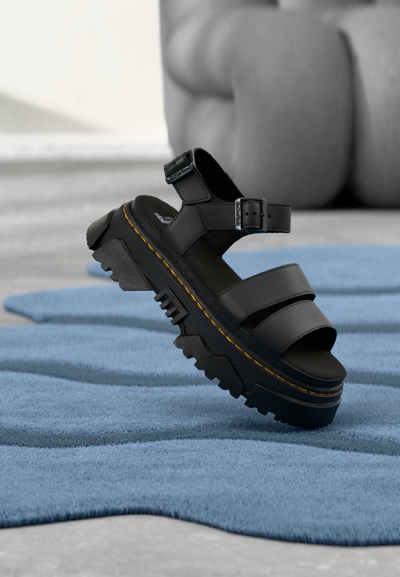 Μαύρο σανδάλι με πλατφόρμα και παχιά σόλα, με ρυθμιζόμενο λουρί στον αστράγαλο και κίτρινη ραφή, τοποθετημένο πάνω σε υφή μπλε υφάσματος.