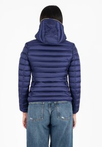 Gewatteerde marineblauwe jas met een capuchon, voorzien van horizontale gestikte lijnen. Het materiaal lijkt glanzend en heeft een gladde textuur. Gecombineerd met lichtblauwe jeans.