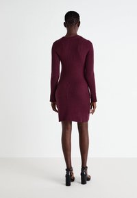 Abito lavorato a maglia di un intenso bordeaux scuro con una texture a coste, maniche lunghe e un design aderente. Indossato con sandali neri a tacco cubo.