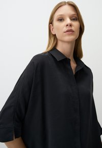 Black, loose-fitting shirt with a collared neckline and short sleeves. Soft texture, features button closure and side slits at the sleeves.