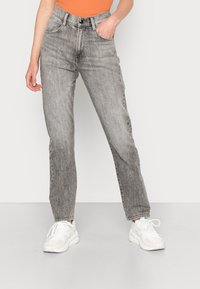 Casaco de ganga cinza com corte reto, apresentando uma cintura alta, desbotamento subtil, bolsos frontais e fecho com botão. Combinado com ténis brancos.