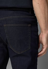 Dunkelblaue Jeans mit geradem Schnitt, sichtbaren Nähten und einer einzelnen Tasche auf der Rückseite. Der Stoff hat eine glatte, leicht verwitterte Textur.