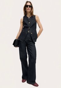 Gilet in denim scuro con bottoni dorati, abbinato a pantaloni a zampa in denim scuro. Accessori comprendono una borsa nera e scarpe rosse con motivo.