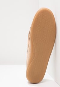 Chaussure à enfiler avec une tige en suède beige clair et une semelle en caoutchouc texturé. La semelle présente un motif ondulé pour une meilleure traction.