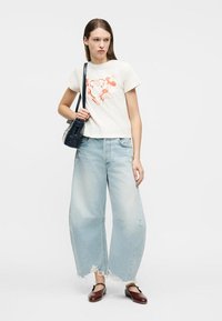 Γυναίκα που φορά λευκό t-shirt με στάμπα Mickey Mouse, ανοιχτό μπλε, φαρδιά, τζιν με σκισίματα, σκούρα τσάντα ώμου και καφέ παπούτσια Mary Jane.