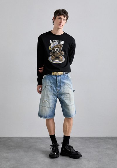Pulover negru tricotat cu grafică de urs, pantaloni scurți din denim albastru cu detalii distruse și pantofi negri cu șireturi. Accent de curea MOSCHINO aurie.