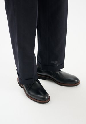 Czarne skórzane buty na formalne okazje z zapięciem na sznurówki, okrągłym noskiem i brązową obszywką; noszone z ciemnymi spodniami w prążki, ukazując elegancki design i gładką fakturę.