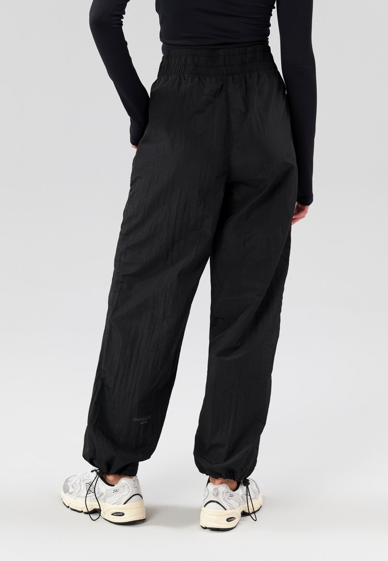 Personne portant un pantalon noir ample avec taille et poignets élastiques, associée à des baskets blanches, debout avec une main dans la poche, vue de dos.