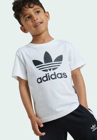 Bela bombažna majica s prenaglašenim črnim adidas logotipom in obliko lista. V kombinaciji s črnimi trenirko in vidnimi žepi.