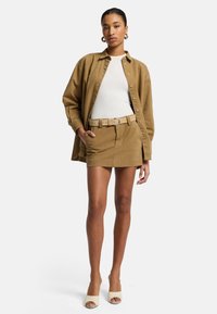Tan oversized button-up shirt over a white fitted top, gecombineerd met een tan minirok met riem en beige open teen hakken. Gladde texturen overal.