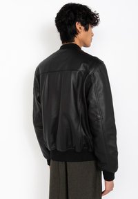 Hombre de perfil vistiendo una chaqueta bomber de cuero negra con puños y cinturilla acanalados, de pie contra un fondo blanco.