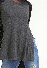 Top gris de manga corta en un material suave con un corte holgado. Presenta una parte trasera de punto abierto en negro y llega hasta las caderas. Combinado con jeans azules desgastados.