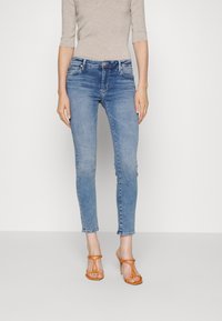 Vaqueros skinny de mezclilla azul claro con cintura media, ligera distorsión y cinco bolsillos; combinados con tacones naranjas de tiras.