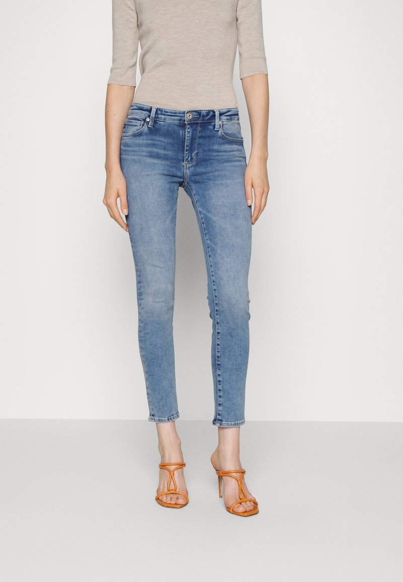 Vaqueros skinny de mezclilla azul claro con cintura media, ligera distorsión y cinco bolsillos; combinados con tacones naranjas de tiras.