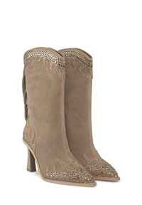 Botines de ante color beige con punteras afiladas, decorados con tachuelas metálicas y patrones de cortes intrincados, que presentan un tacón apilado.