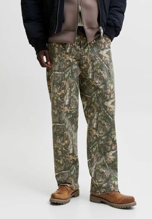 Homme portant un pantalon camouflage, des bottes de travail marron, une veste sombre et un sweat à capuche marron zippé, les mains dans les poches sur un fond uni.