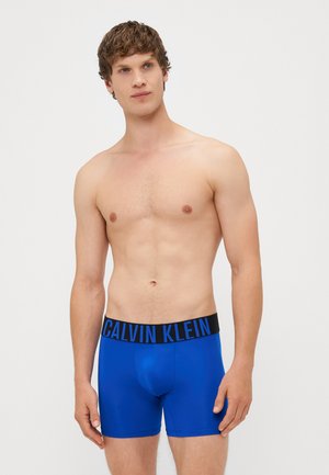 Modri bokserice s črnim elastičnim pasom, na katerem je v modri barvi napis "CALVIN KLEIN". Gladka textura in prilegajoča zasnova.