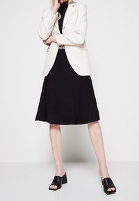 Blazer bianco sopra un dolcevita nero e una gonna nera, abbinato a mules neri aperti. Il tessuto appare liscio e ben rifinito.