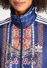 Marinblå zip-up-jacka med hög krage, prydd med intrikata blommiga och geometriska mönster, broderade detaljer och tre vita ränder på axlarna.