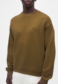 Personne à la peau foncée portant un sweat-shirt vert olive uni à col rond avec un petit logo crocodile brodé sur la poitrine.