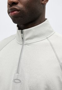 Κοντινό πλάνο ατόμου που φοράει ανοιχτό γκρι πουλόβερ με φερμουάρ μέχρι τη μέση, με μικρό λογότυπο κάτω από το φερμουάρ στο στήθος.