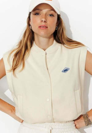 Veste sans manches crème avec un col haut, boutons-pression à l'avant et logo brodé bleu. Texture lisse ; dispose de poches latérales.