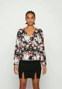 Top blusado floral com mangas longas, fundo preto, flores rosa e brancas, decote em V, usado com uma saia preta justa com uma fenda frontal.
