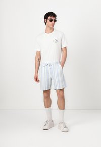 T-shirt blanche avec logo, short de bain à rayures bleu clair et blanches, baskets blanches et lunettes de soleil. Le modèle se tient devant un fond neutre.