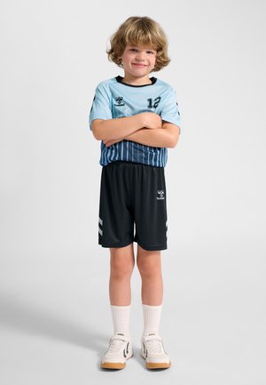 Ein lächelnder junger Junge mit blondem Haar steht mit verschränkten Armen, trägt hellblaue und schwarze Sportbekleidung, weiße Socken und weiße Sneakers.