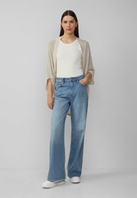 Hellbeige offene Strickjacke über einem weißen Tanktop, kombiniert mit hochgeschnittenen hellblauen Schlagjeans und weißen Sneakers vor einem grauen Hintergrund.