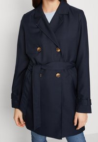 Trench coat azul marino de doble pecho con cinturón en la cintura, hecho de una tela suave. Presenta botones en tono dorado, bolsillos laterales y puños enrollados.