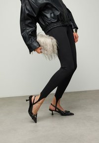 Veste aviateur en cuir noir avec taille élastique, leggings noirs ajustés et escarpins noirs à brides arrière avec bouts pointus et boucle décorative.