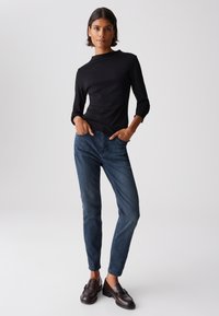 Schwarzes Langarmoberteil, taillierte Passform, Stehkragen. Blaue Denim-Jeans, schmaler Schnitt, leichte Ausbleichung. Braune Slipper. Neutraler Hintergrund.