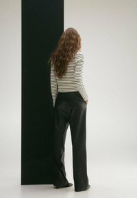 Schwarze Leder-Hosen mit weitem Bein, kombiniert mit einem figurbetonten, gestreiften Langarmoberteil aus glattem Material. Das Model steht vor einer vertikalen, schwarzen Wand.