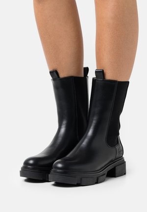 Widok od nóg osoby noszącej czarne skórzane buty do połowy łydki z masywną podeszwą i pętelkami do zakładania, stojącej na białej powierzchni.